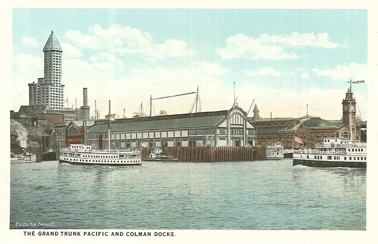 Carte postale colorisée des entrepôts du port de Seattle, avec deux bateaux à vapeur amarrés, des grues et la tour pointue du Smith Tower au fond.