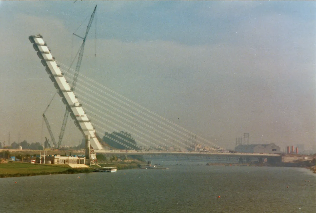 Ponte do Alamillo em construção sobre o rio, com mastro inclinado, cabos, guindastes e estruturas metálicas na margem.