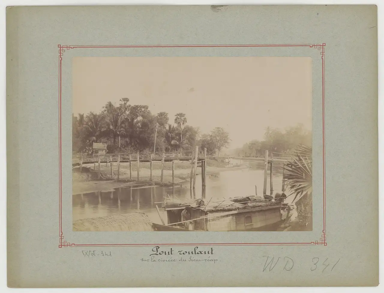 Uma ponte de madeira estreita atravessa o rio Siem Reap, com um barco coberto de palha ancorado abaixo e palmeiras na margem.