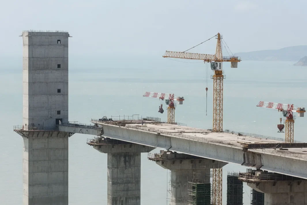 O viaduto em obra sobre a água, com gruas nos vãos e a torre sem letreiro à esquerda.