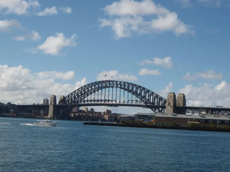 Um arco de aço sobre a água, com a cidade baixa atrás e um barco ao longe.