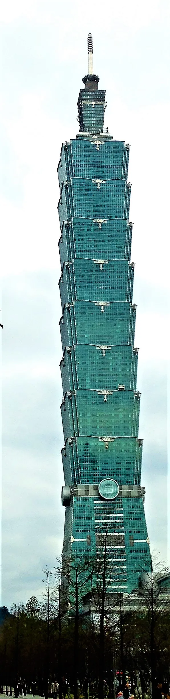 A torre de vidro em degraus, com a antena no topo, vista entre árvores.