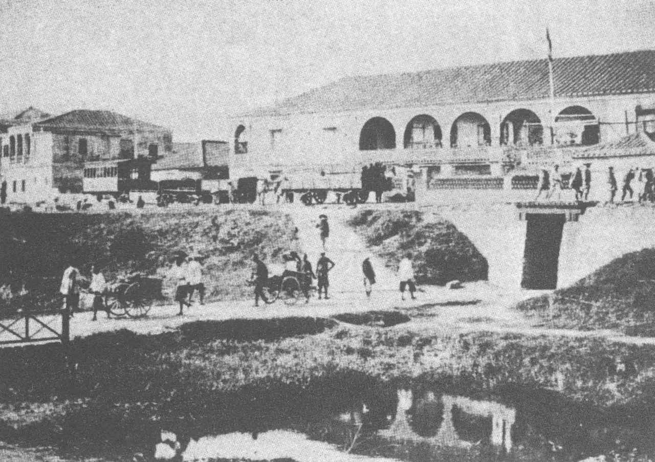 Fotografia da estação de Taipé, um edifício baixo com arcadas e telhado de telhas, com vagões, riquixás e pessoas a pé num caminho de terra diante de um canal.