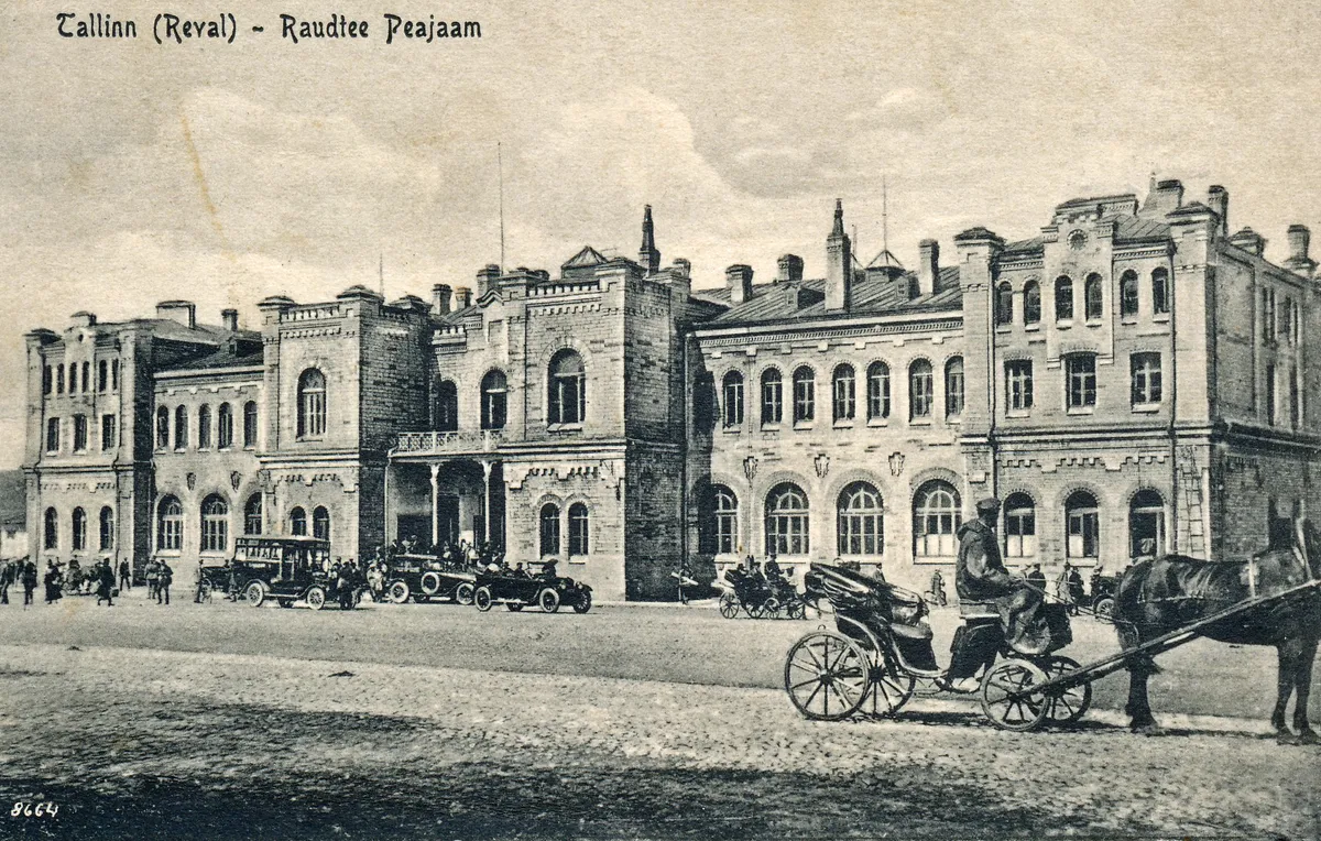 Postal com título da estação, edifício de pedra com chaminés e janelas em arco, automóveis, carruagens, cavalo e pequenas figuras.