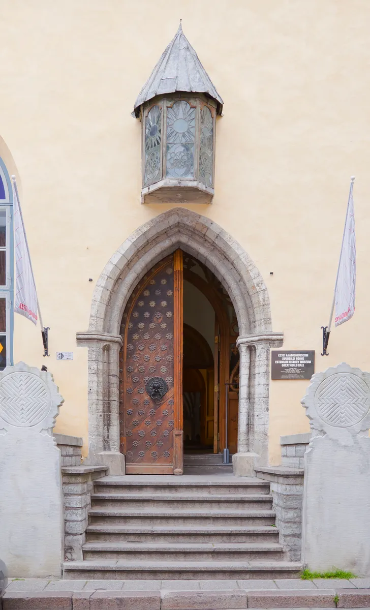 Portal de pedra em arco, porta de madeira entreaberta com ornamentos de metal, escadaria, vitral superior e bandeiras laterais.