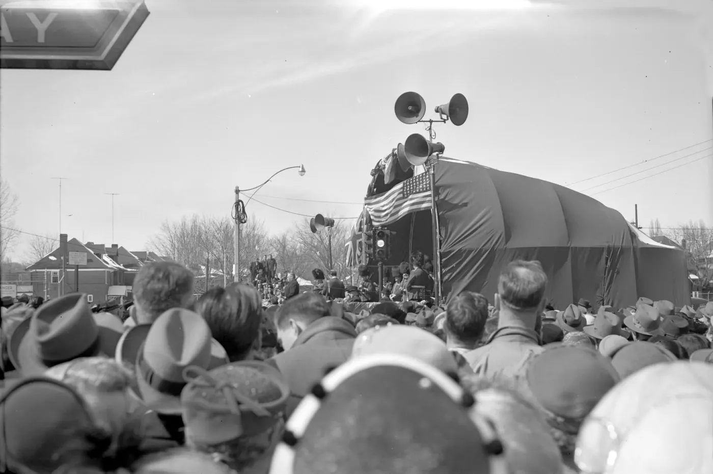 Uma multidão de costas diante de um palco com bandeiras, na frente de uma estação.