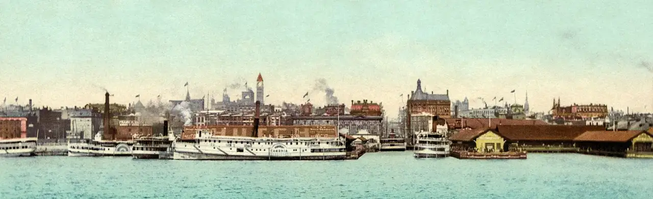 Postal en fotocromo de la costa de Toronto vista desde la bahía, con barcos de vapor amarrados en los muelles y detrás edificios de ladrillo, una torre de reloj y chimeneas.