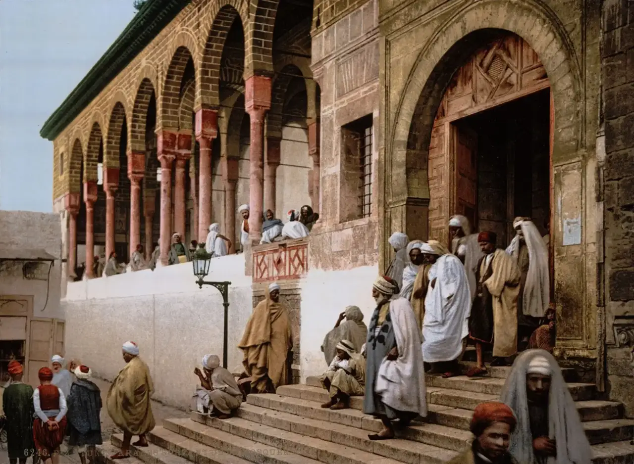 Fotocromo de homens de túnica e turbante nos degraus de uma mesquita de Túnis, com uma porta em arco de madeira e uma galeria de colunas vermelhas no alto.
