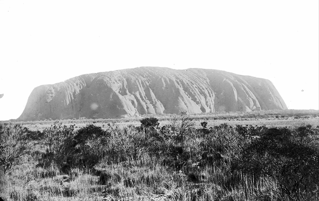 Fotografia em preto e branco do Uluru, o monólito de encostas sulcadas, visto de longe sobre uma planície com arbustos e capim no primeiro plano.