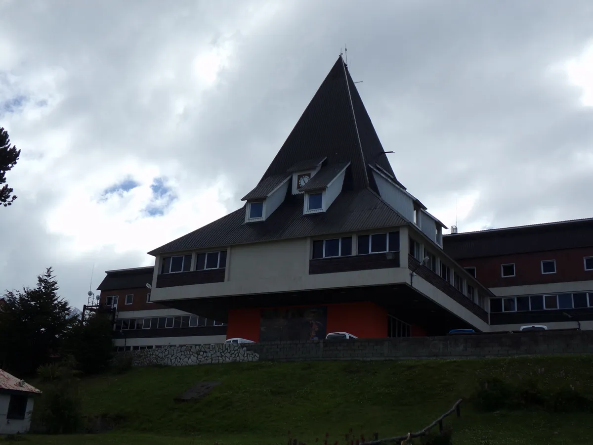 Edifício com telhado triangular escuro e um relógio na água-furtada, sobre um volume laranja, num gramado.