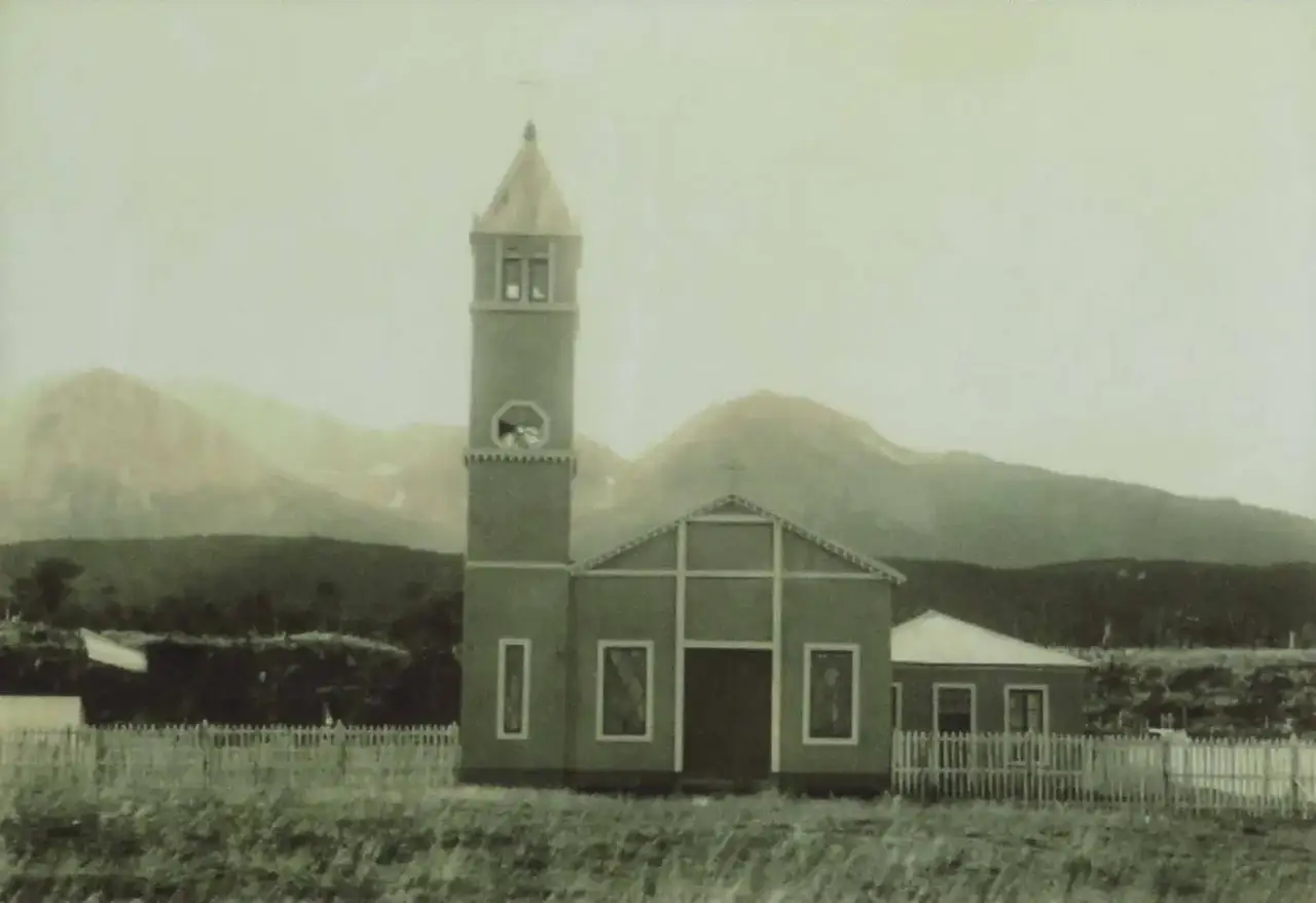 Fotografia da primeira igreja católica de Ushuaia, com torre de relógio e cerca de madeira branca, diante de montanhas cobertas de mato.