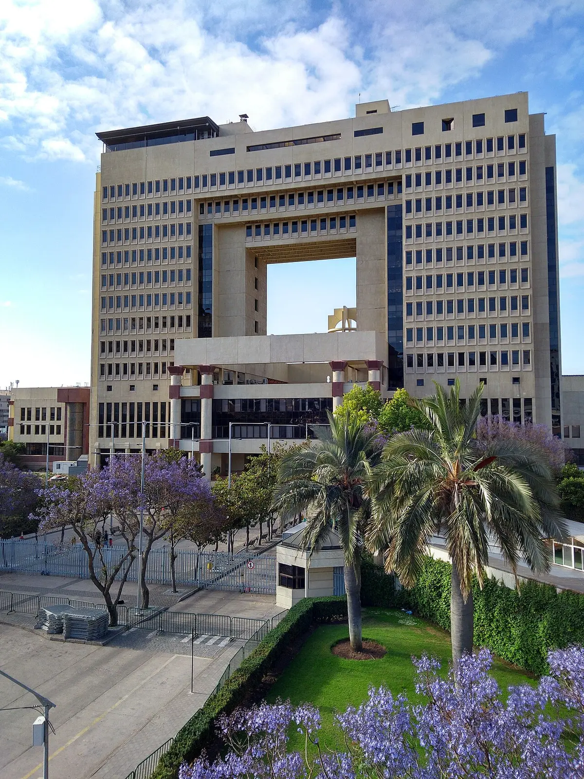 Torre bege com um vão retangular no centro, palmeiras e árvores de flor roxa na frente, céu azul.