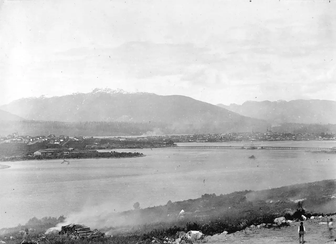 Fotografia de False Creek vista do sul, com a ponte de madeira da Cambie Street cruzando a água, a cidade baixa na outra margem e montanhas ao fundo.