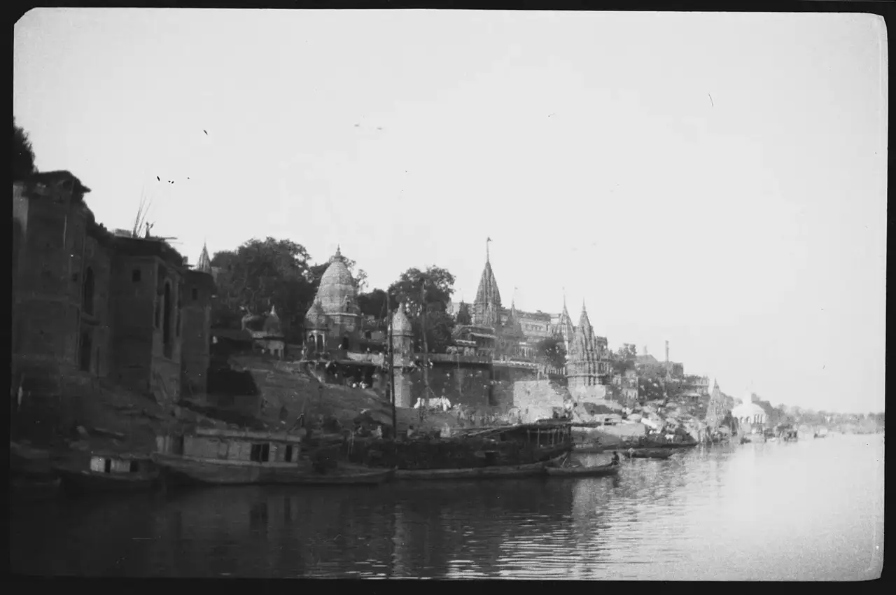Fotografia tirada do rio Ganges, com barcos na margem e templos de torres pontiagudas sobre os ghats, na direção do Manikarnika Ghat.
