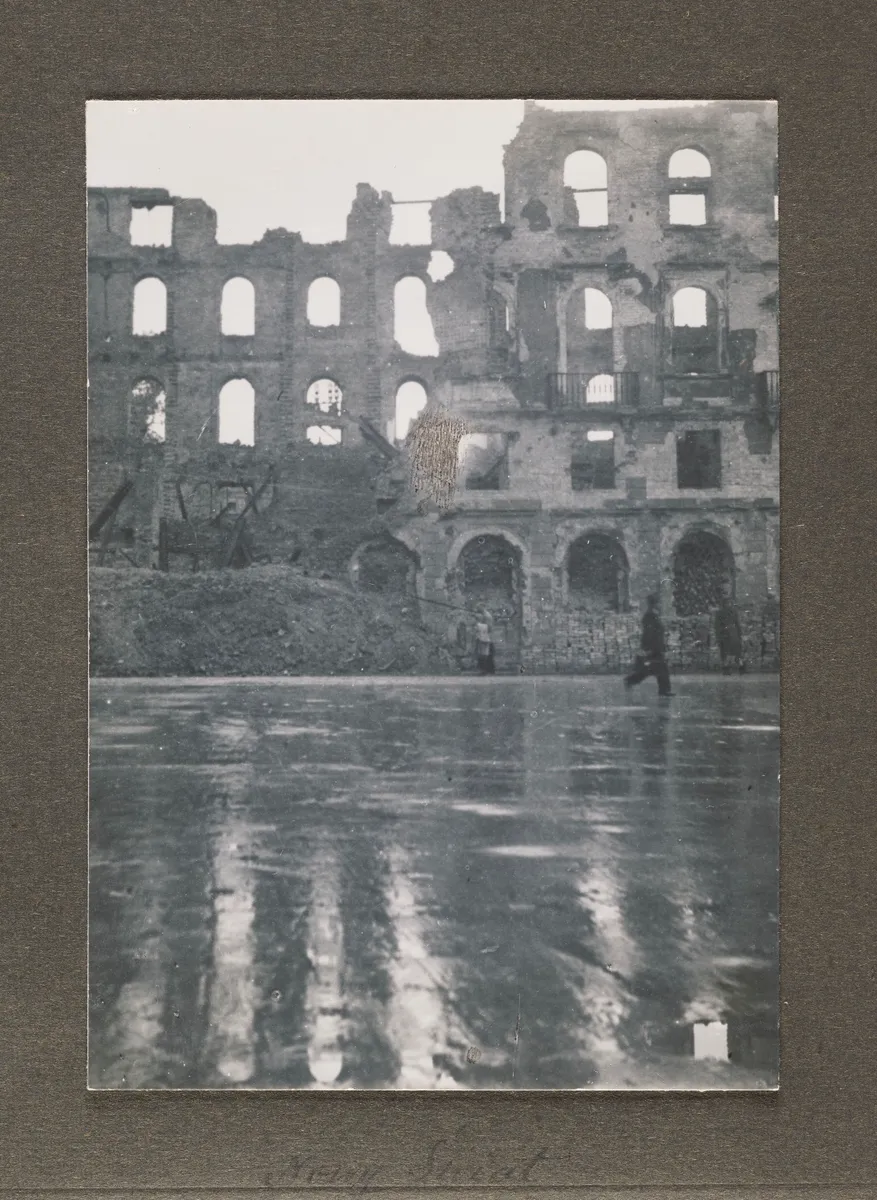 Fotografia em preto e branco numa folha de álbum escura, com fachadas em ruínas, janelas vazias, poça refletindo os edifícios e pequenas figuras na rua.