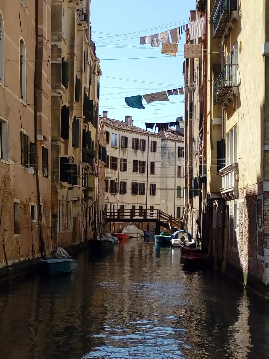 Canal estreito entre edifícios antigos de tijolo e reboco, com ponte de madeira, roupa estendida e barcos amarrados.