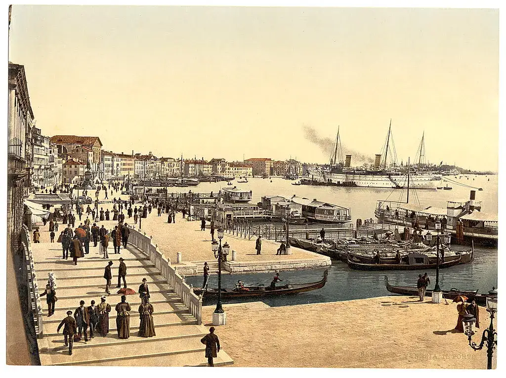 Fotocromo da orla de Veneza com uma ponte de degraus cheia de pedestres, gôndolas atracadas, barcos a vapor e um navio de mastros e chaminés na laguna.