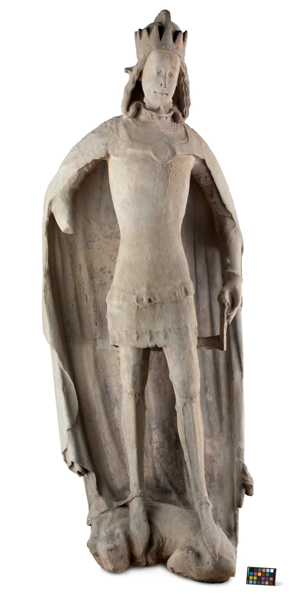 Medieval sculpture of the crowned Rudolf IV, standing with a cloak and animals at the base, photographed against a pale background.