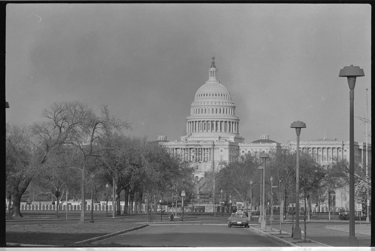 Photographie en noir et blanc du Capitole au fond d’une rue avec voiture, arbres et lampadaires, ciel gris et marges sombres et claires du négatif.