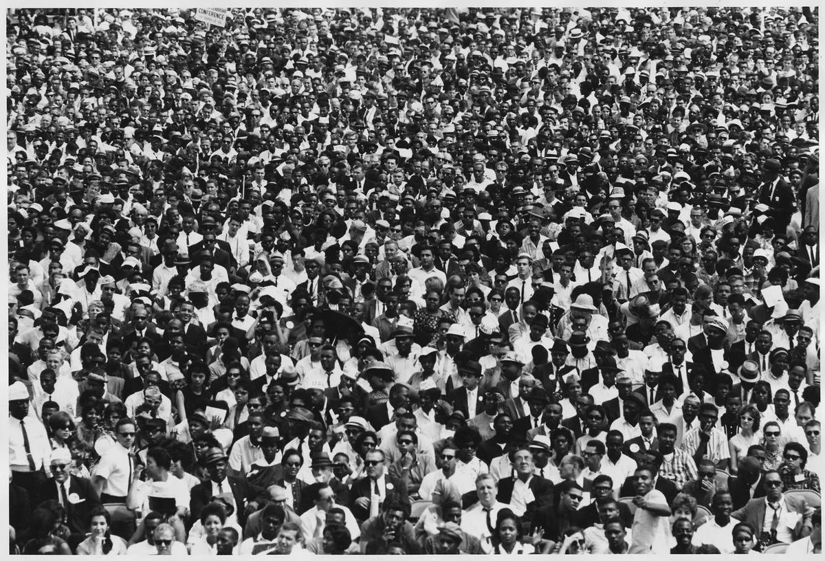 Fotografia em preto e branco de uma multidão reunida, com pessoas usando chapéus e óculos e alguns cartazes ao fundo, dentro das bordas claras da imagem.