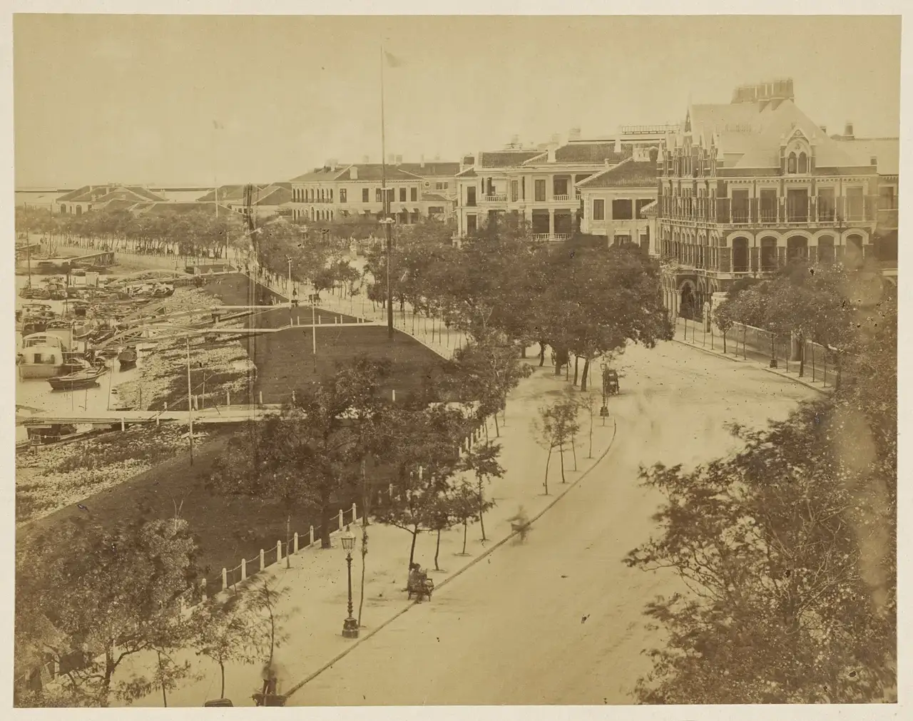 Fotografia vista do alto do Bund de Xangai, com uma avenida curva arborizada, um gramado à beira do rio com barcos e sobrados de varanda, entre eles o consulado holandês.