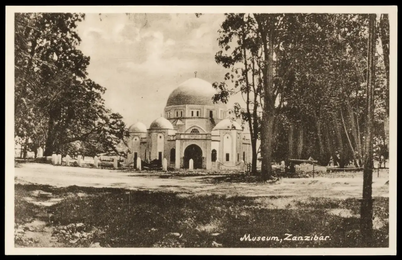 Cartão-postal com a legenda Museum, Zanzibar: um edifício de cúpulas brancas entre árvores altas, com lápides baixas à esquerda e um terreno aberto à frente.